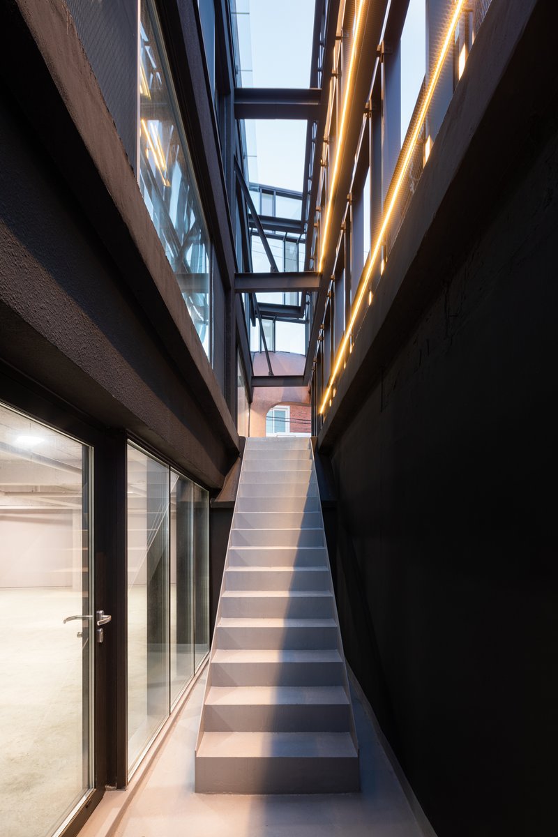 Narrow interior stair passage between black walls with steel-framed bridges and warm strip lighting at dusk