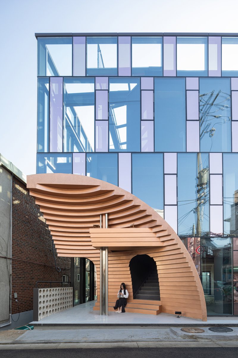 Curved timber-slat pavilion entrance with a person descending, set against a blue glass facade