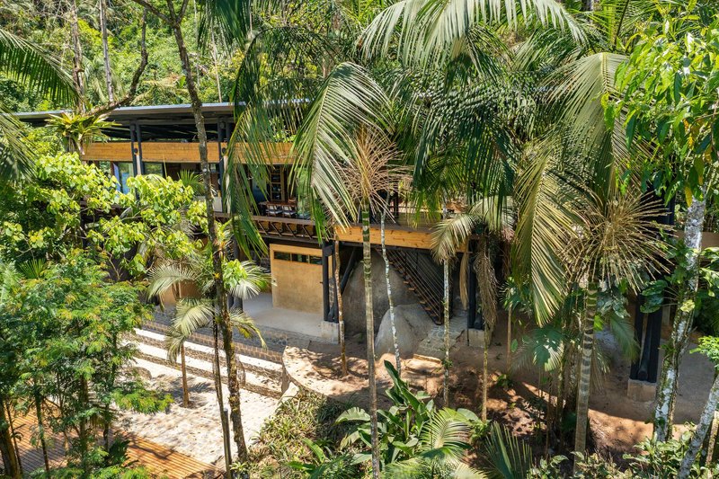 Two-story volume with timber and rammed earth walls framed by palm fronds and jungle vegetation