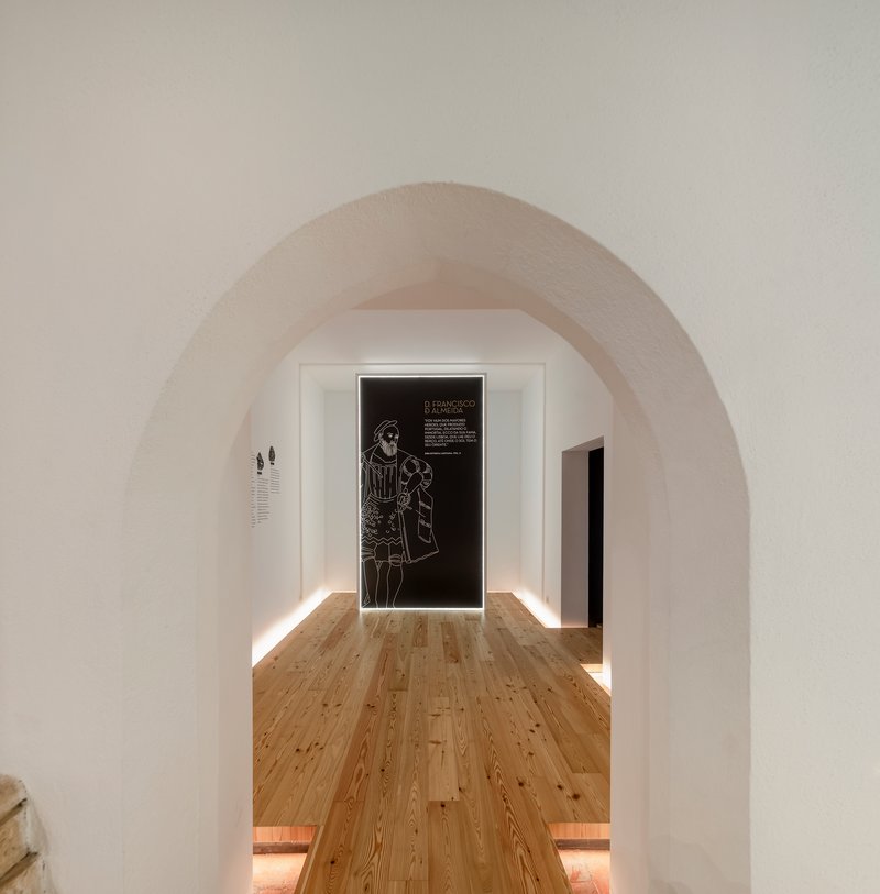 View through whitewashed stone arch toward gallery space with backlit graphic panel on far wall