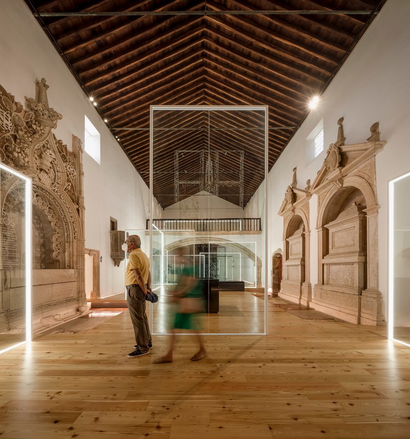 Visitors standing near glass display box in a gallery with carved stone tombs and timber roof