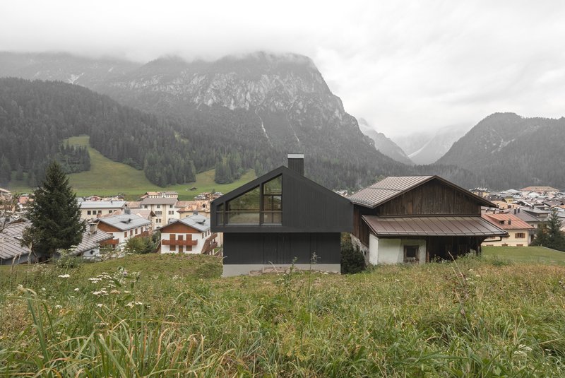 Two-part house with dark cladding and glass volume beside weathered barn under cloudy mountain sky