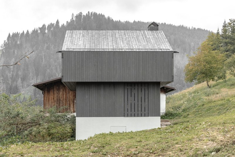 Two-story timber clad volume on a grassy slope with misty forested mountains in the background