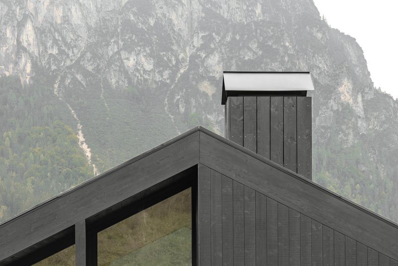 Vertical chimney clad in dark timber rising above the gabled roofline against a rocky mountain face