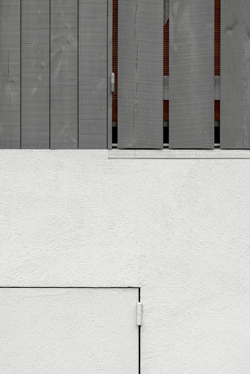 Concrete facade with vertical timber-lined recesses above a white stucco wall and paved courtyard