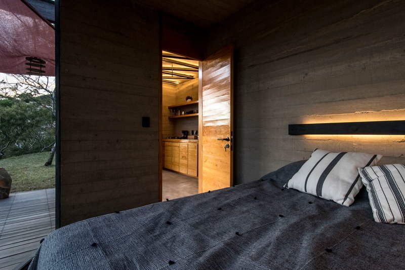 Bedroom with concrete walls and recessed lighting looking through open door to timber kitchenette