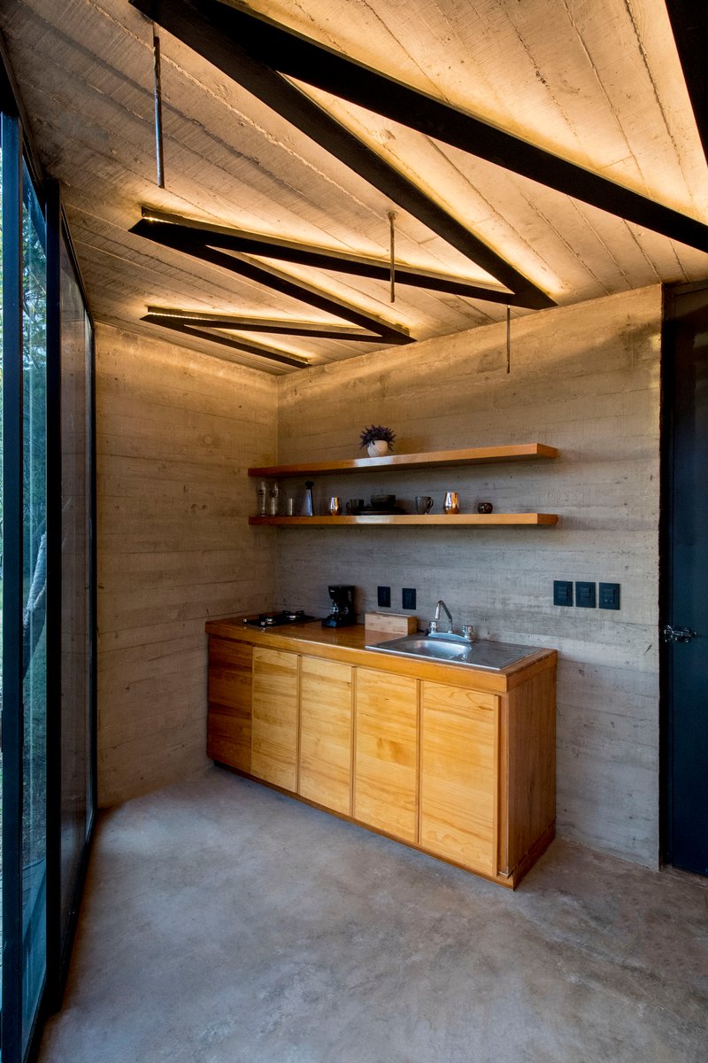 Compact kitchen with timber cabinetry beneath triangular suspended light fixtures and board-formed concrete walls