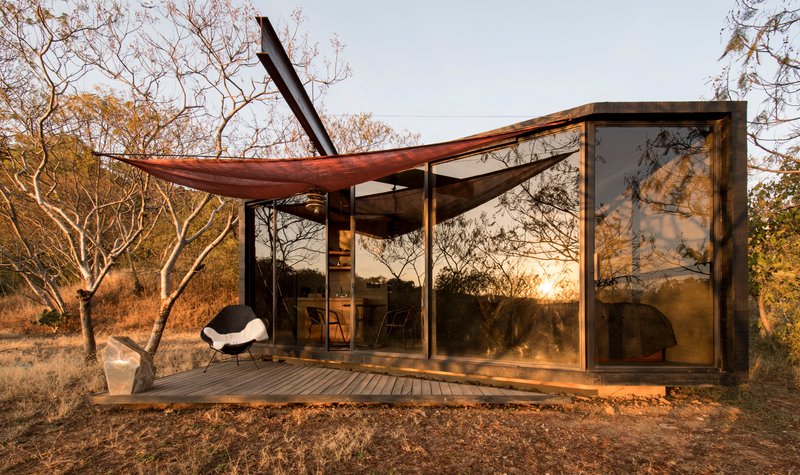 Glazed facade with deck and fabric shade canopy among bare winter trees at dusk