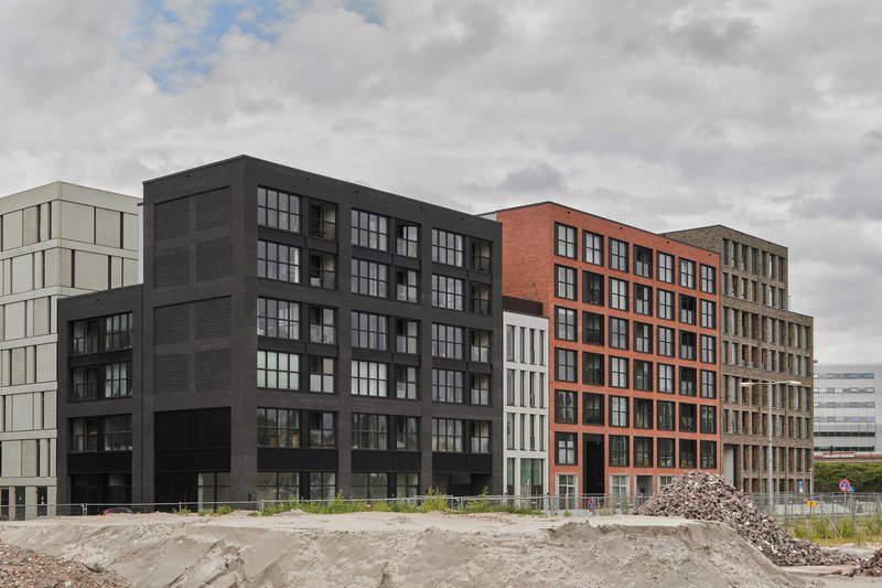 Street view of three multiunit blocks with orange brick, black cladding and grey panel facades across undeveloped ground