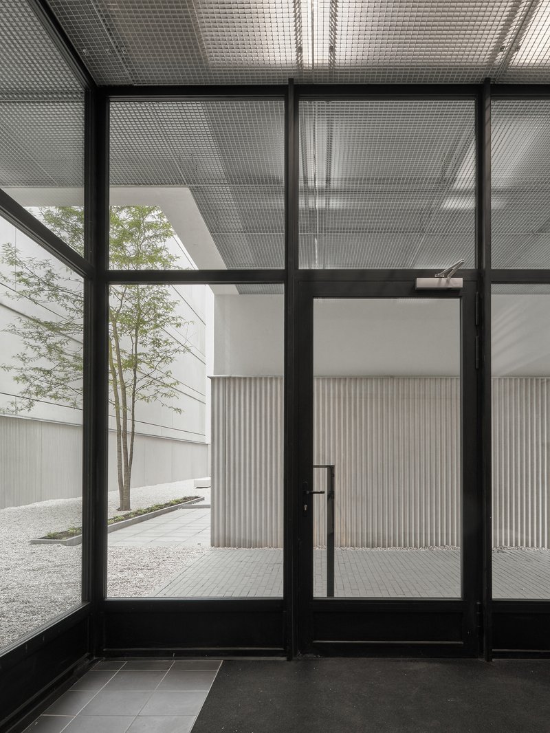Black-framed glass entry doors with translucent corrugated overhead canopy and gravel courtyard beyond