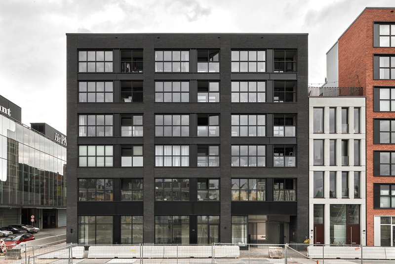 Black cladded facade with large gridded windows and recessed balconies flanked by neighboring buildings