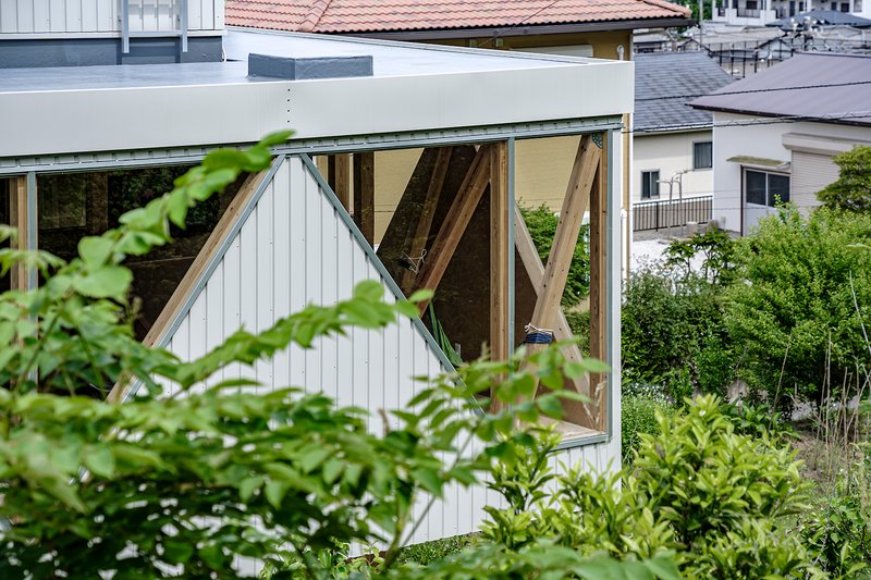 White corrugated metal facade with exposed diagonal timber bracing visible through a glazed opening surrounded by vegetation