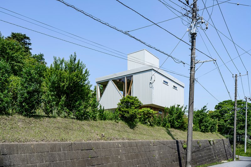 White corrugated metal house on a grassy slope with overhead power lines and concrete retaining wall