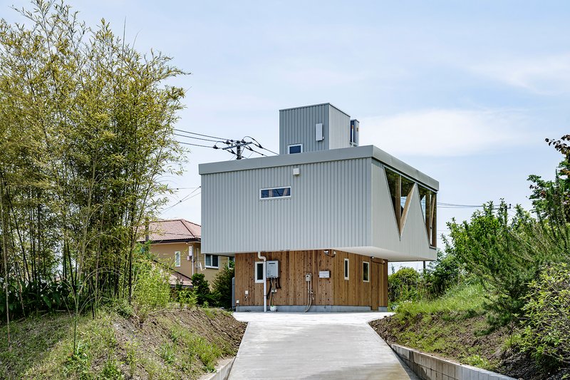 Front facade with corrugated metal upper volume cantilevering over timber-clad ground floor and concrete drive