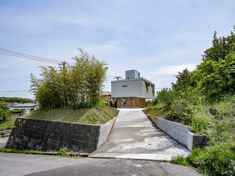 Concrete driveway rising toward a metal-clad house nestled between a wooded hillside and neighboring rooftops