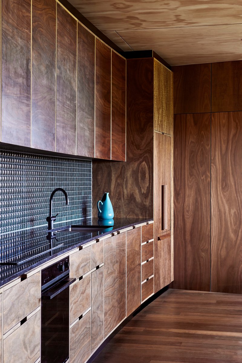 Galley kitchen with textured black tile backsplash and timber cabinetry under plywood ceiling