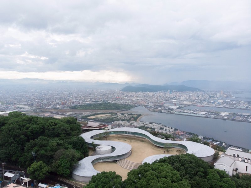 Aerial view of the spiraling white roof nestled among forested hillside overlooking a coastal harbor
