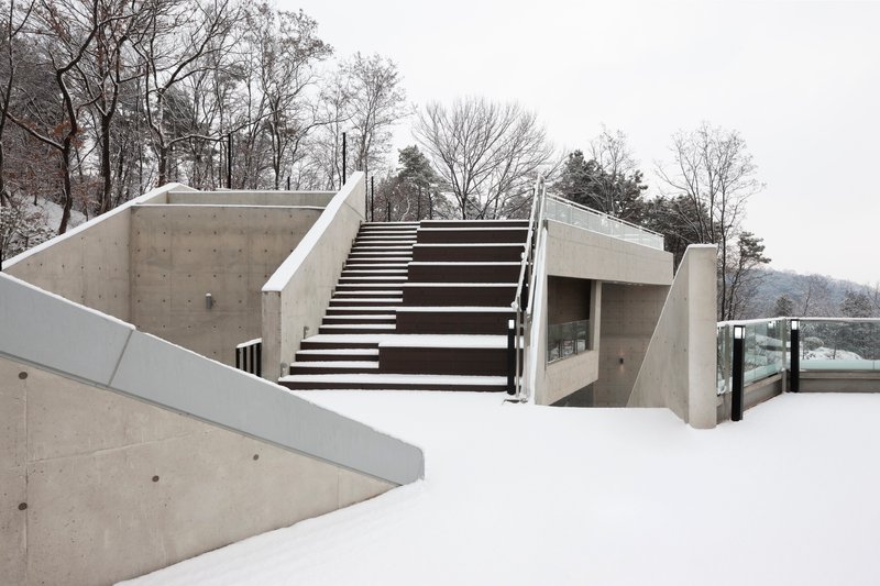 Exterior concrete stair ascending from a snow-covered terrace with bare winter trees in the background