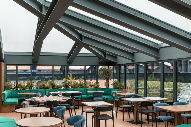 Dining area beneath exposed steel roof trusses with skylights and turquoise banquette seating