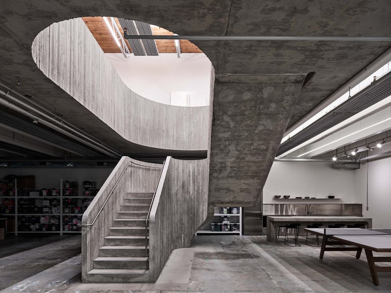 Curved concrete staircase with board-formed walls rising through an oval ceiling opening to natural light