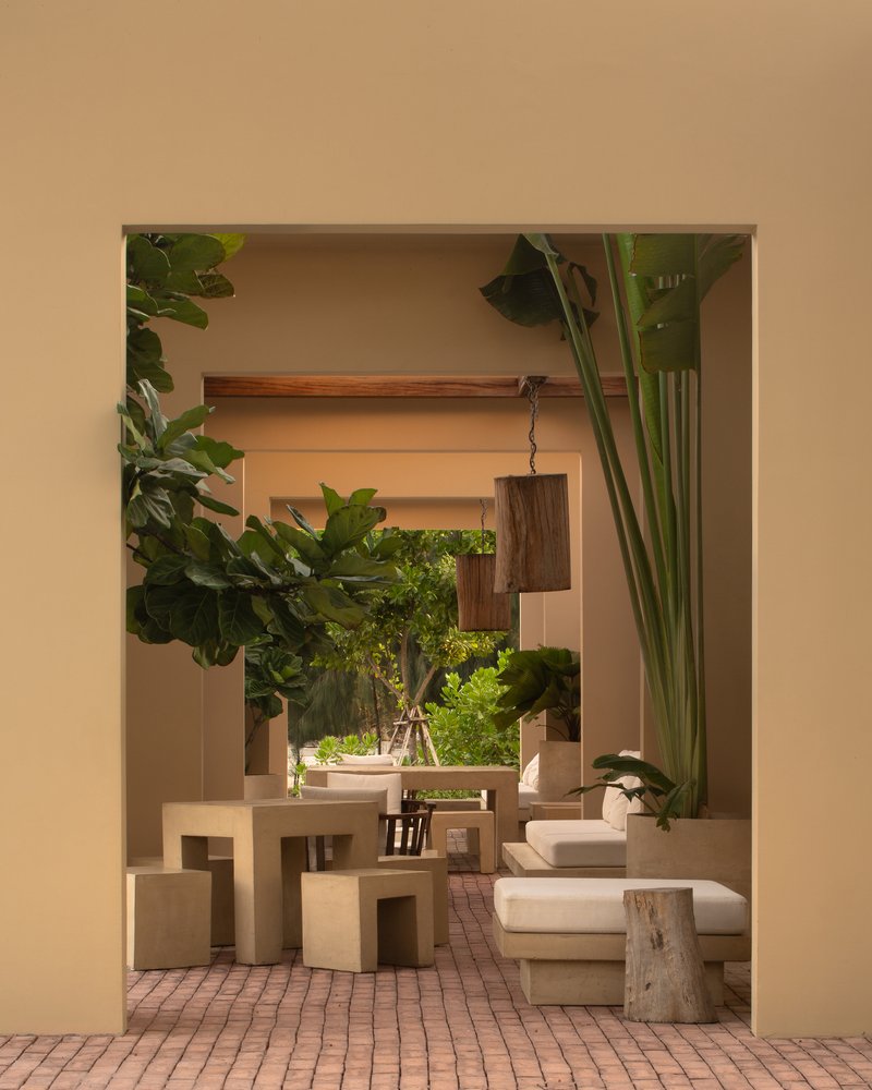 Framed view through an opening showing dining furniture, hanging timber lights, and potted plants on brick pavers
