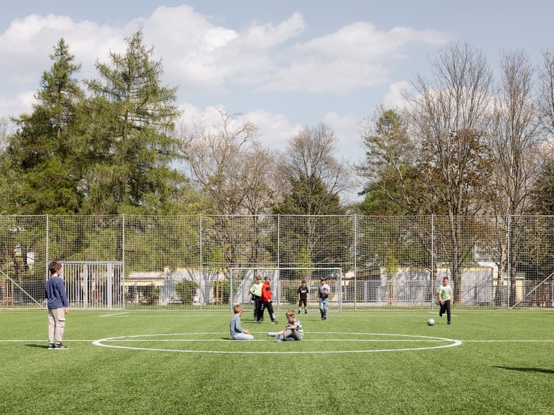 Green artificial turf pitch with mesh fencing and children playing under a partly cloudy spring sky