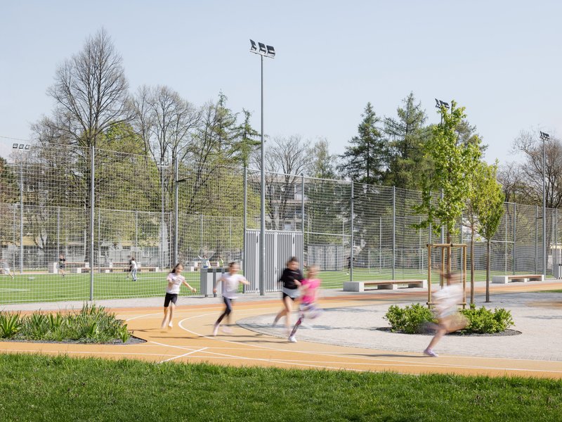 Children running across a curved plaza with planted beds and mesh sports enclosure beyond