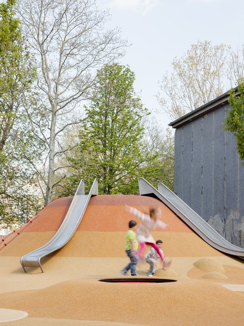 Contoured play mound with stainless steel slides and children running near concrete structure under cloudy sky