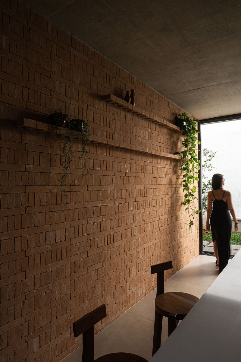Patterned brick wall with timber shelving and trailing vines, a woman walking toward sunlit courtyard