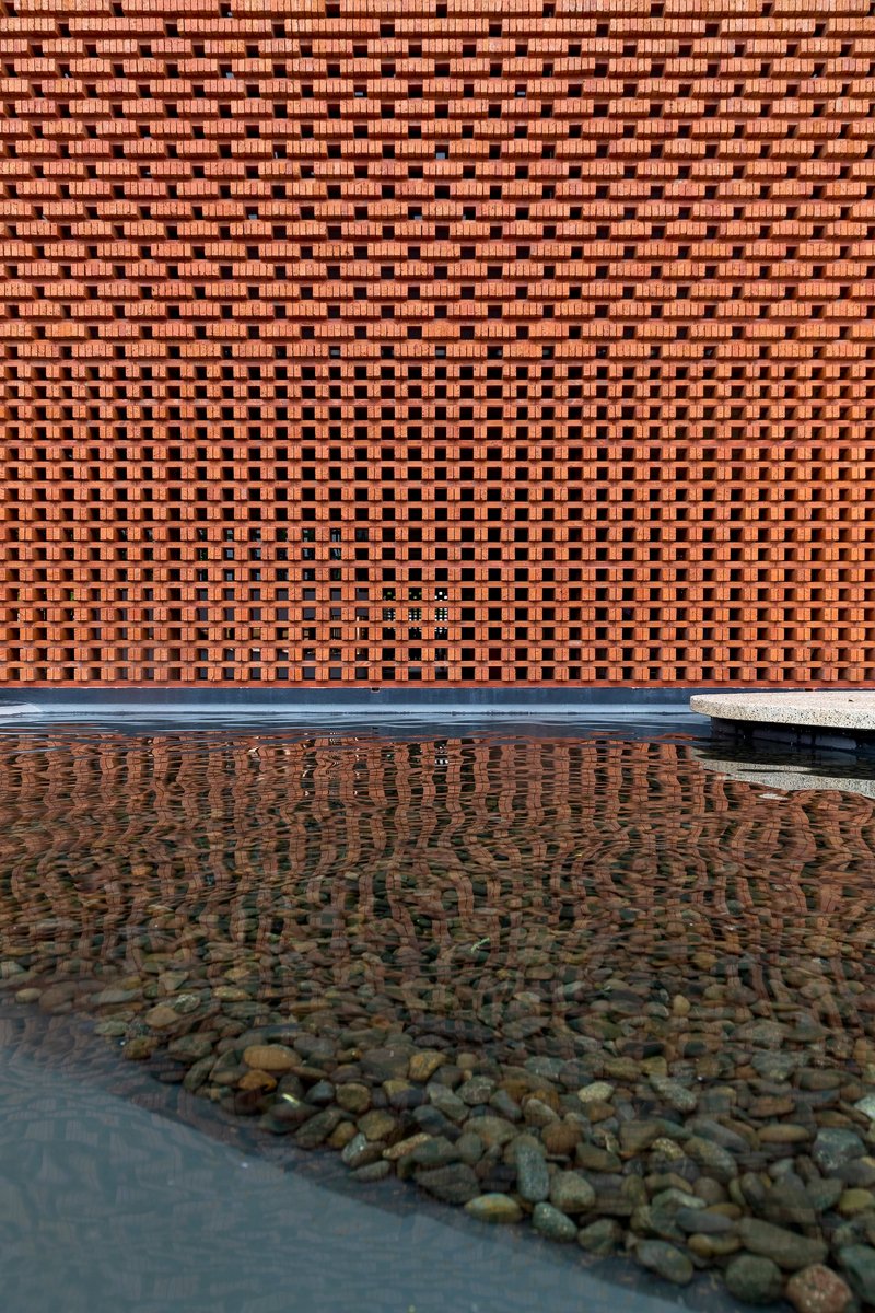 Perforated brick screen wall with varying aperture patterns reflected in shallow water pool with river stones