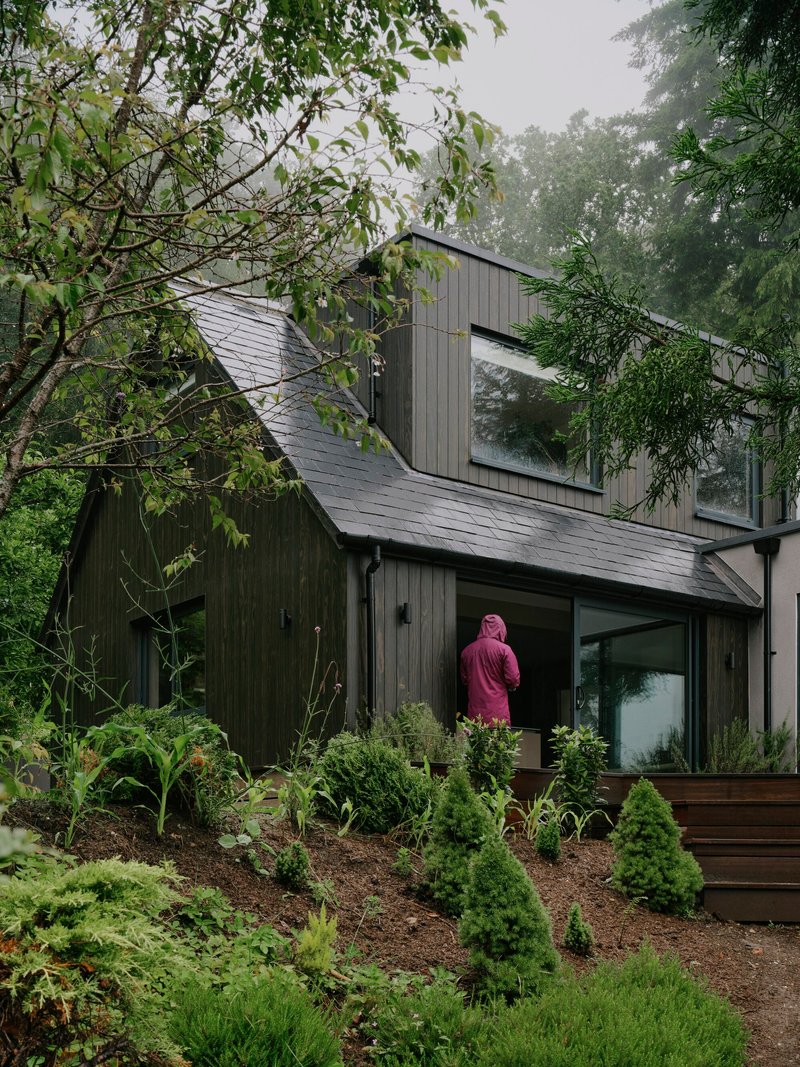 Figure in pink robe on planted slope below the dark timber and slate facade