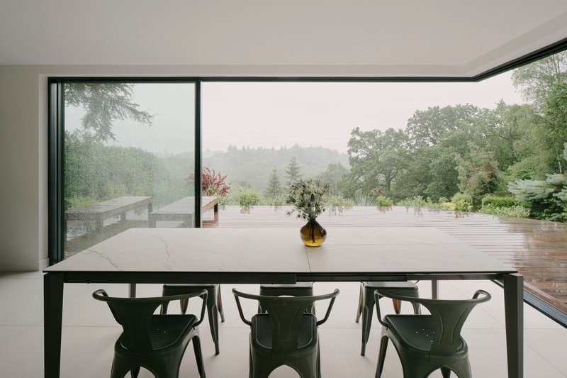 Dining table with marble top and dark chairs framing floor-to-ceiling glazing overlooking a misty landscape