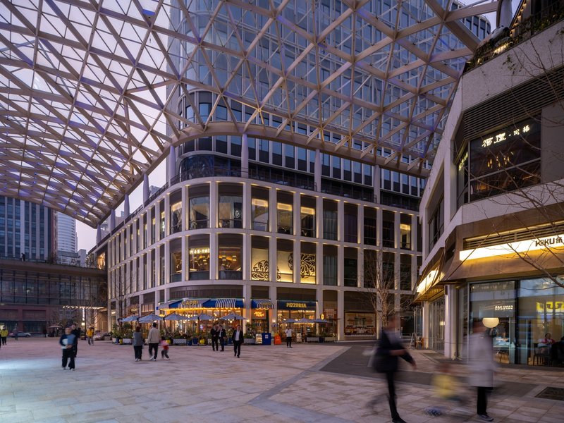 Covered plaza with curved glazed volume and pedestrians moving beneath the diagrid canopy at dusk