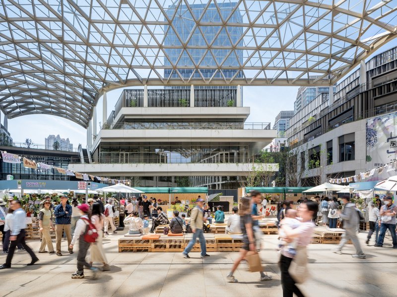 Covered plaza with diagonal lattice canopy sheltering crowds at wooden bench seating below terraced retail facades