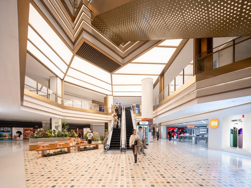 Double-height atrium with chevron-patterned skylight above an escalator and geometric tile flooring with shoppers passing through