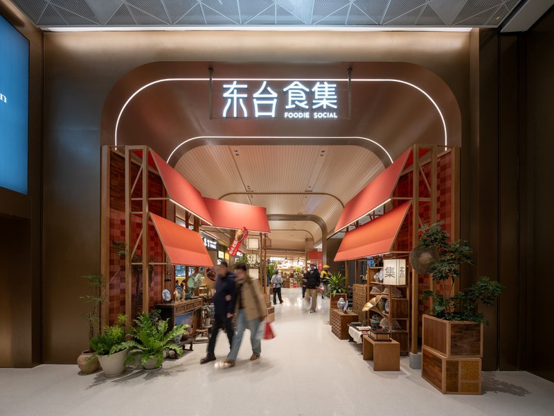 Entrance portal with red fabric banners and timber slat ceiling framing the food market beyond