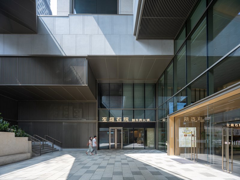 Courtyard entry with vertical metal screening, glass facades, and two figures crossing the paved ground plane