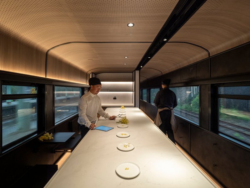 Galley counter with perforated timber ceiling where two chefs prepare dishes while the train is in motion
