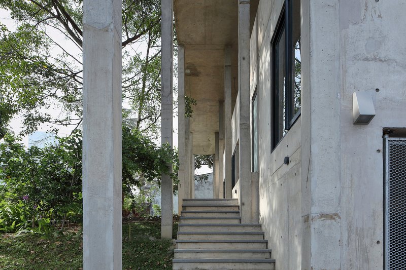 Exterior concrete stair rising between cylindrical columns toward a timber-clad soffit with trees beyond