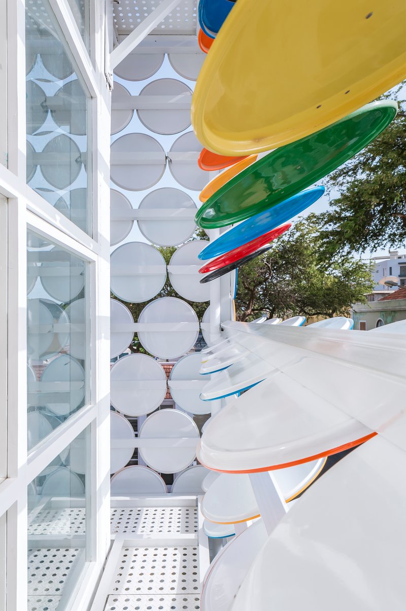 Exterior view of stacked multicolored surfboards against circular glass panel facade and perforated metal deck