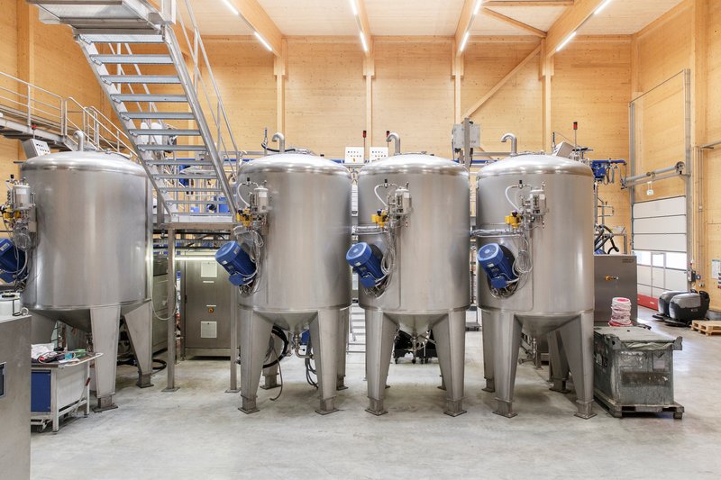 Row of stainless steel fermentation tanks with blue valves on a concrete floor beneath exposed timber beams