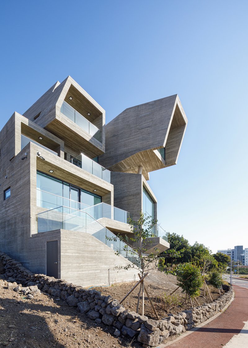 Angular concrete volumes cantilevering over a landscaped slope with young trees and stone edging
