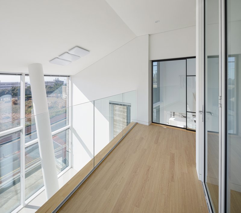 Upper level corridor with timber flooring and glass balustrade overlooking a double-height space flooded with daylight
