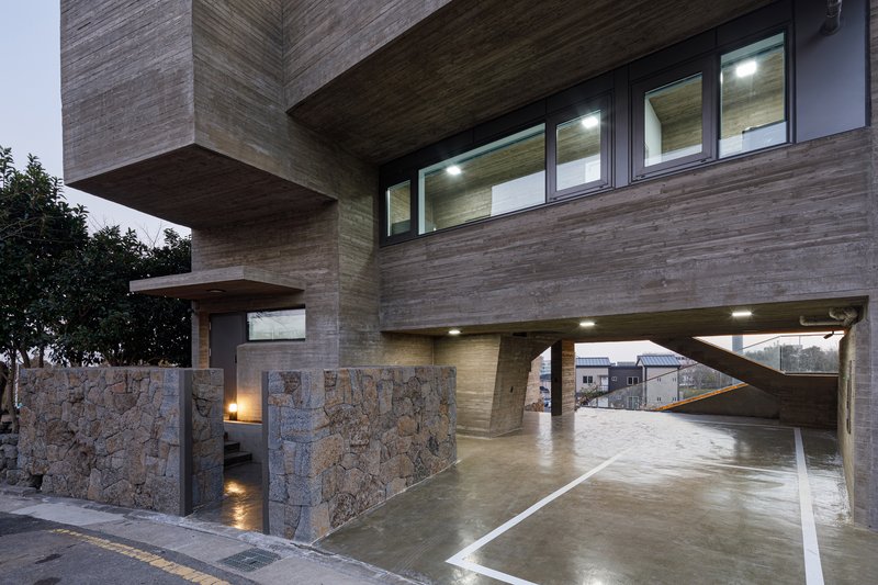 Horizontal timber-clad facade on pilotis above stone-clad retaining walls and wet concrete driveway at dusk
