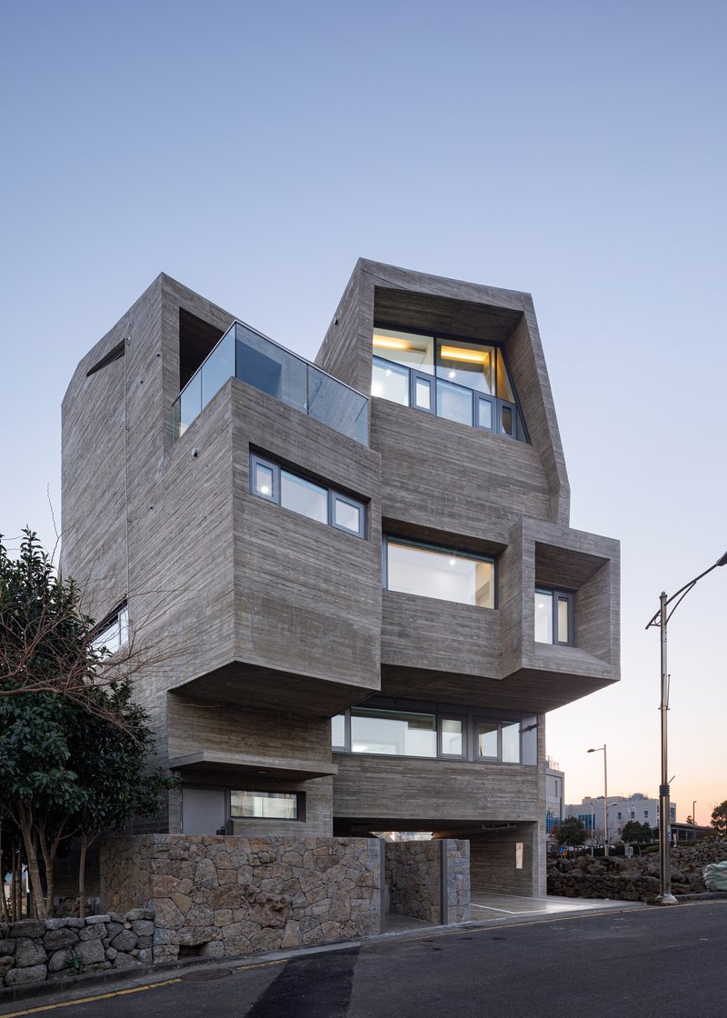 Stacked timber-clad volumes with cantilevered floors and illuminated interiors at twilight beside a residential street