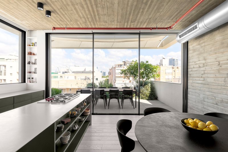 Open kitchen with green cabinetry looking toward glazed terrace doors framed by exposed concrete ceiling and conduit