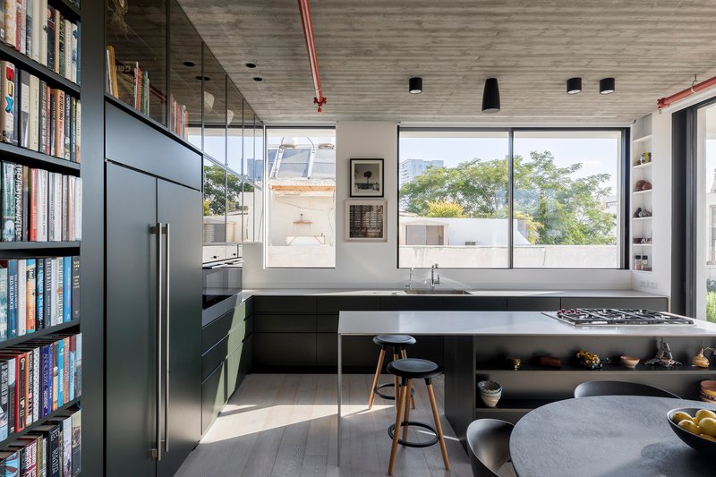 Kitchen island with integrated cooktop beside full-height glazed partition reflecting trees outside in afternoon light