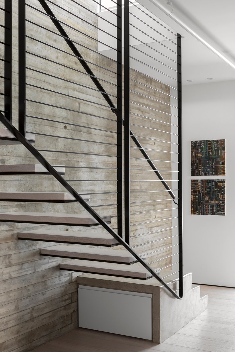 Steel floating staircase with horizontal cable railing against board-formed concrete wall under a skylight