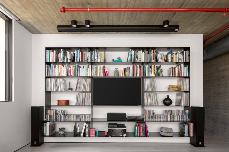 Built-in black shelving unit with television and books below exposed concrete ceiling with red conduit