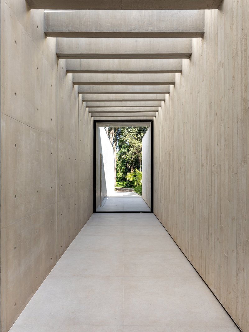 Concrete corridor with exposed formwork and repeating transverse beams framing a doorway to greenery beyond